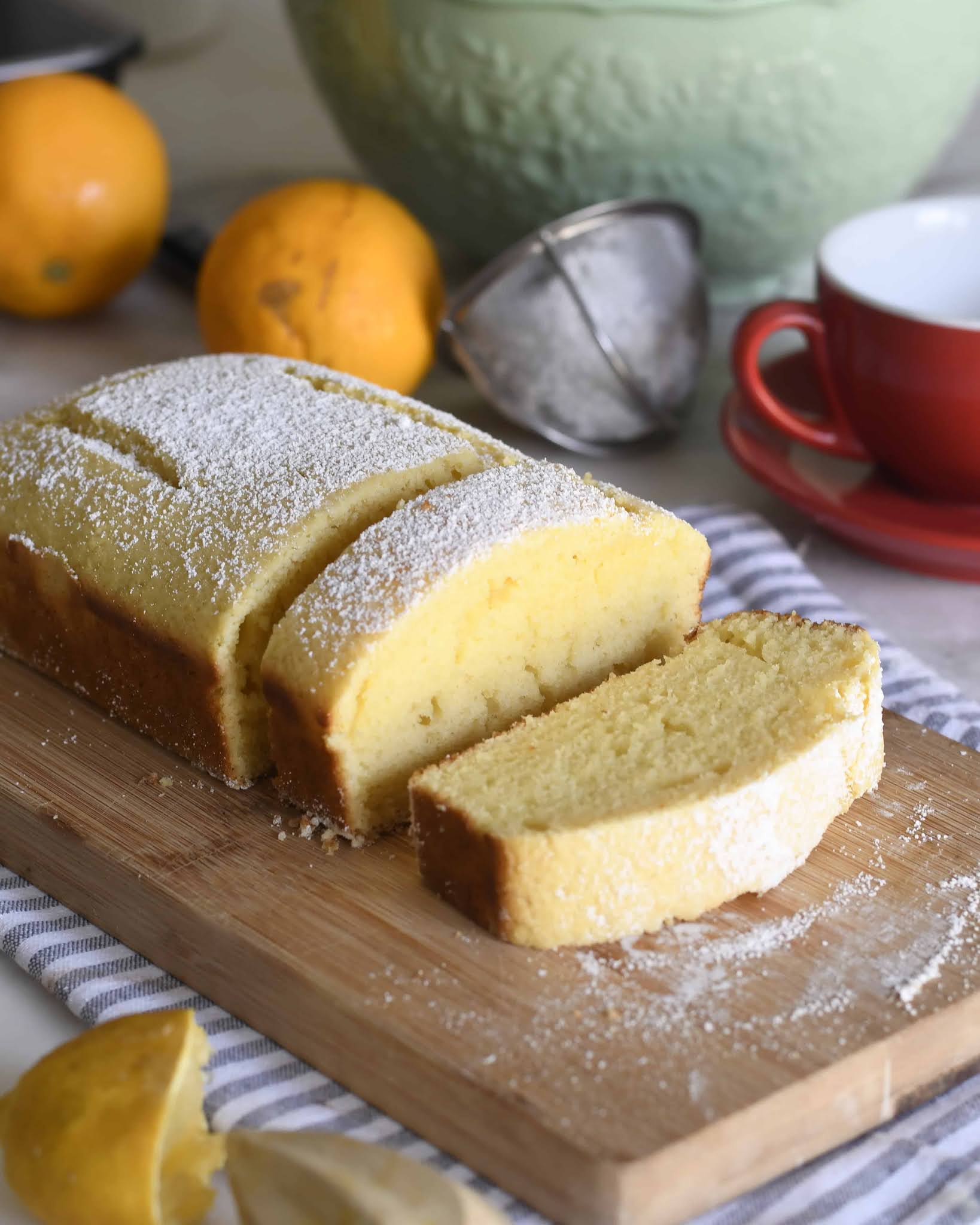 A loaf of lemon cake dusted with powdered sugar sits on a wooden cutting board, with one slice cut. Nearby are fresh lemons, a green mixing bowl, a red cup and saucer, and a citrus juicer.