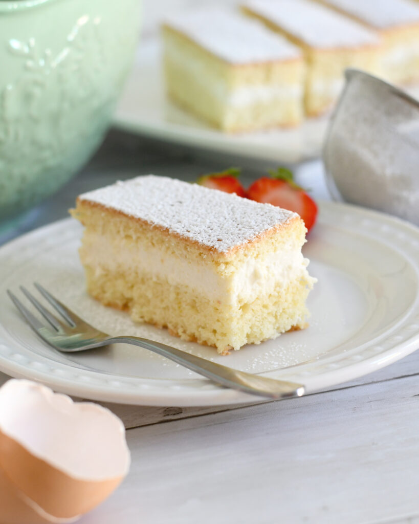 A slice of sponge cake with cream filling and powdered sugar on top sits on a white plate with a fork. Two strawberries and a broken eggshell are nearby, with more cake pieces and a mixing bowl in the background.