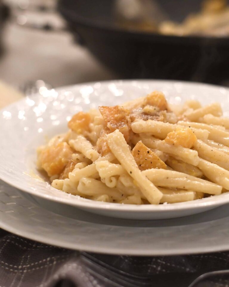 A close-up of a white plate filled with creamy pasta and chunks of golden squash, topped with a sprinkle of black pepper. The background is softly blurred, focusing attention on the dish.