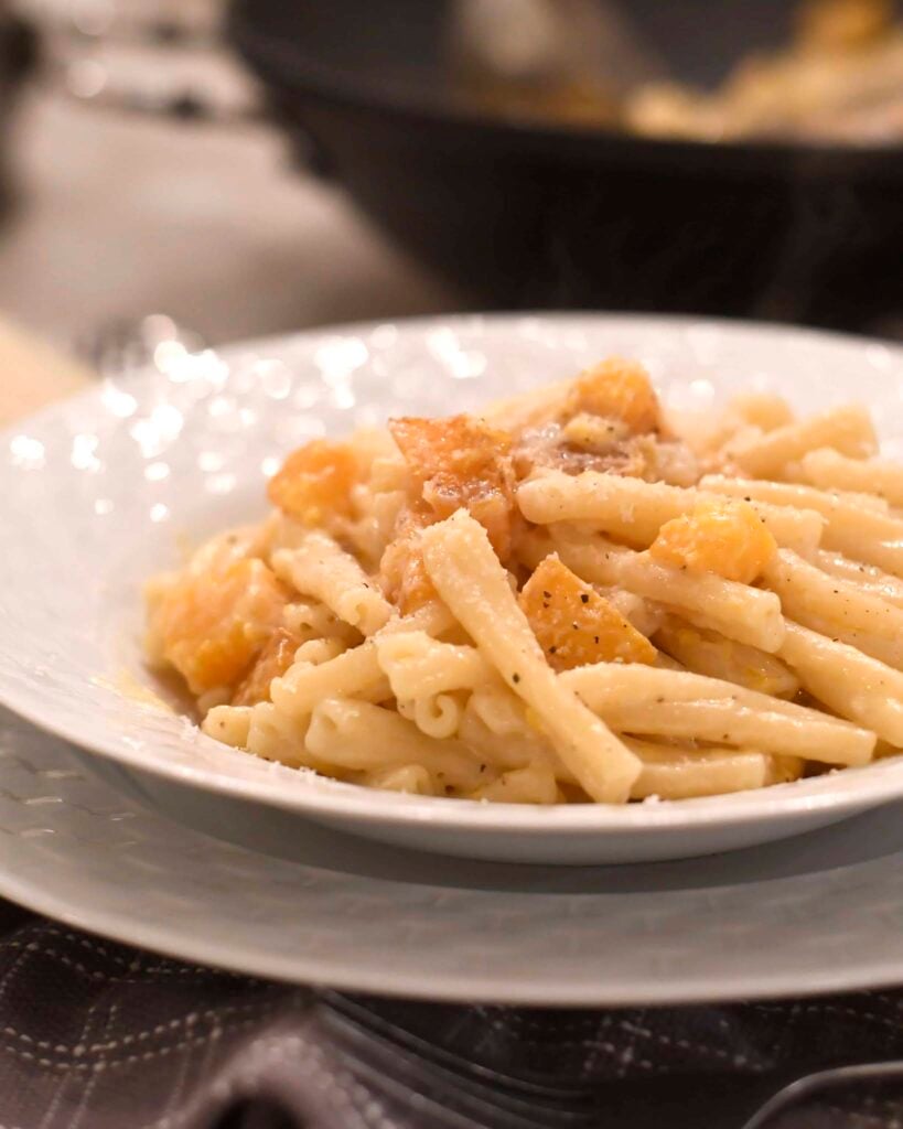 A close-up of a white plate filled with creamy pasta and chunks of golden squash, topped with a sprinkle of black pepper. The background is softly blurred, focusing attention on the dish.