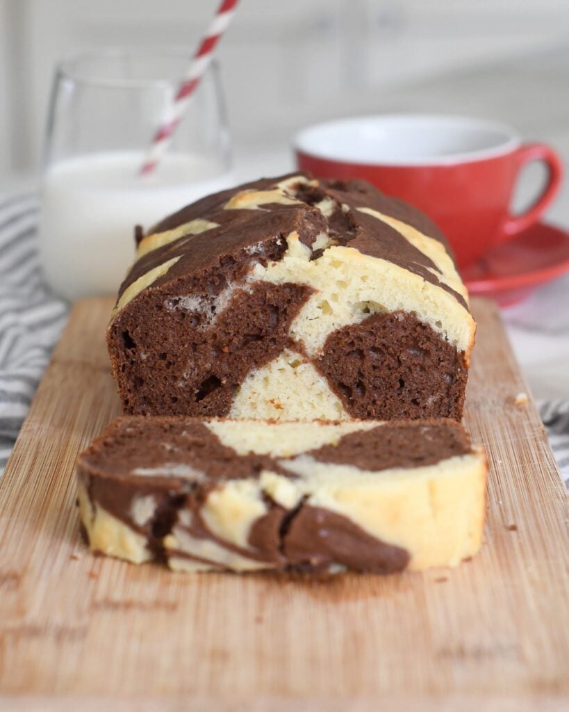A sliced marble cake with chocolate and vanilla swirls sits on a wooden board. Behind it are a glass of milk with a red striped straw and a red cup and saucer. The background is softly blurred.