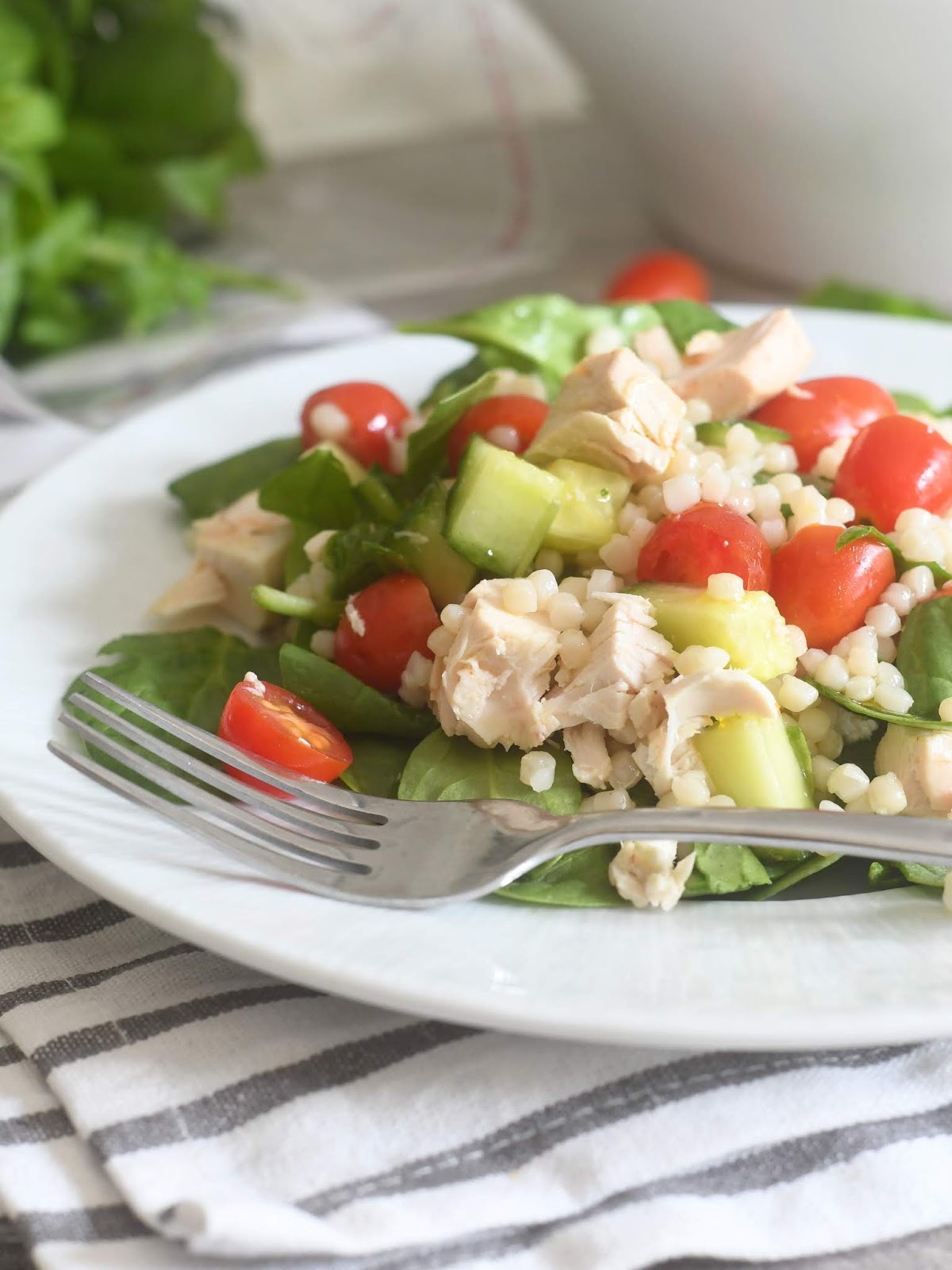 A plate of salad with cherry tomatoes, diced chicken, couscous, cucumber, and greens, with a fork placed on the plate. The setting includes a striped napkin and blurred leafy greens in the background.