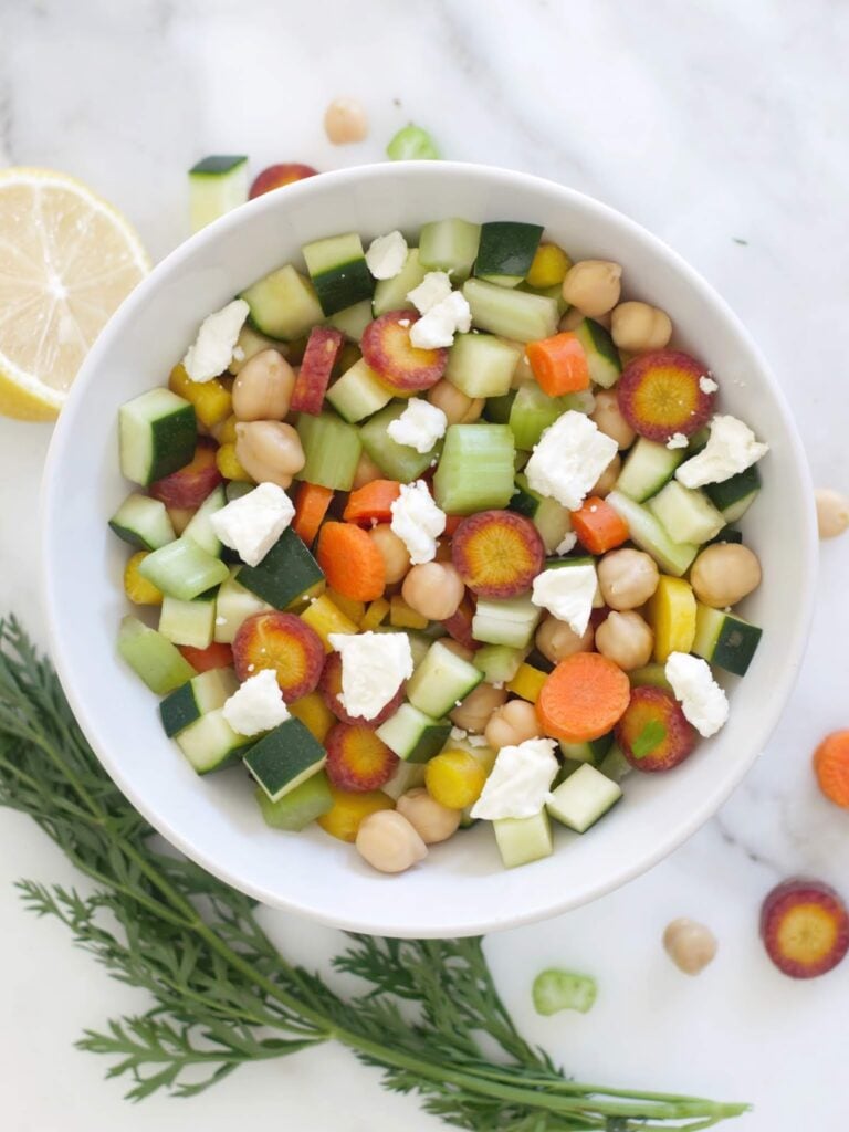 A white bowl filled with a salad of sliced colorful carrots, cucumber, chickpeas, and crumbled white cheese, set on a marble surface with a lemon wedge and carrot greens nearby.