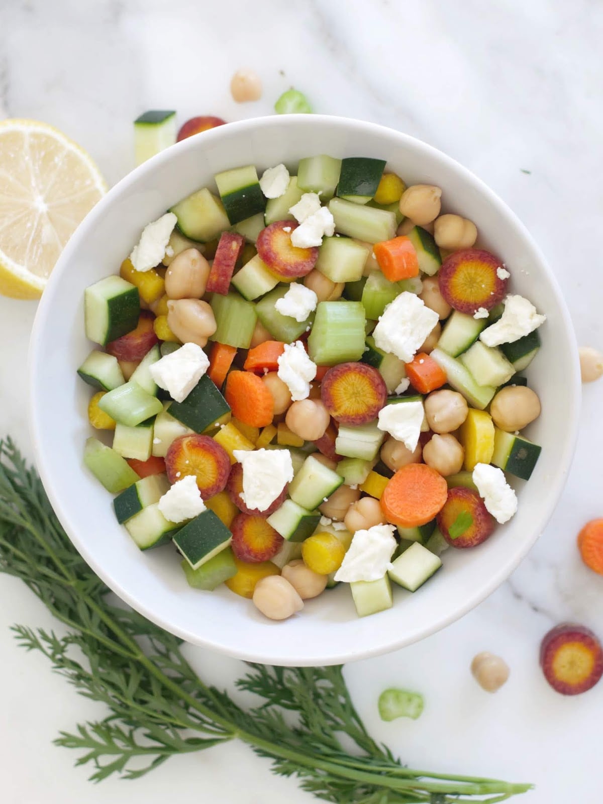 A white bowl filled with a salad of sliced colorful carrots, cucumber, chickpeas, and crumbled white cheese, set on a marble surface with a lemon wedge and carrot greens nearby.