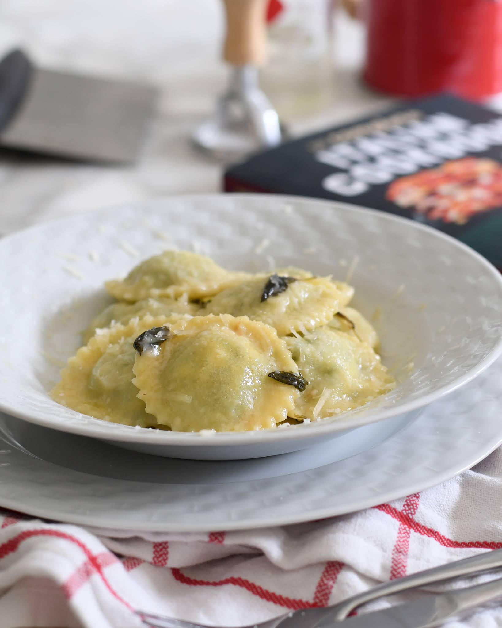 A white bowl filled with spinach ravioli in a light sauce, garnished with herbs, sits on a plate atop a white and red striped cloth. Kitchen utensils and a packaged food box are blurred in the background.