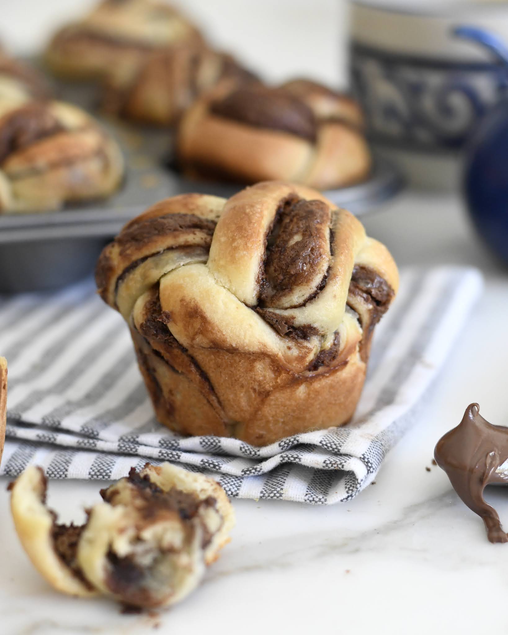 A muffin-shaped babka with chocolate swirls sits on a striped cloth, with a torn piece showing its soft texture. In the background are more babka muffins, a blue patterned mug, and a spoon with chocolate spread.