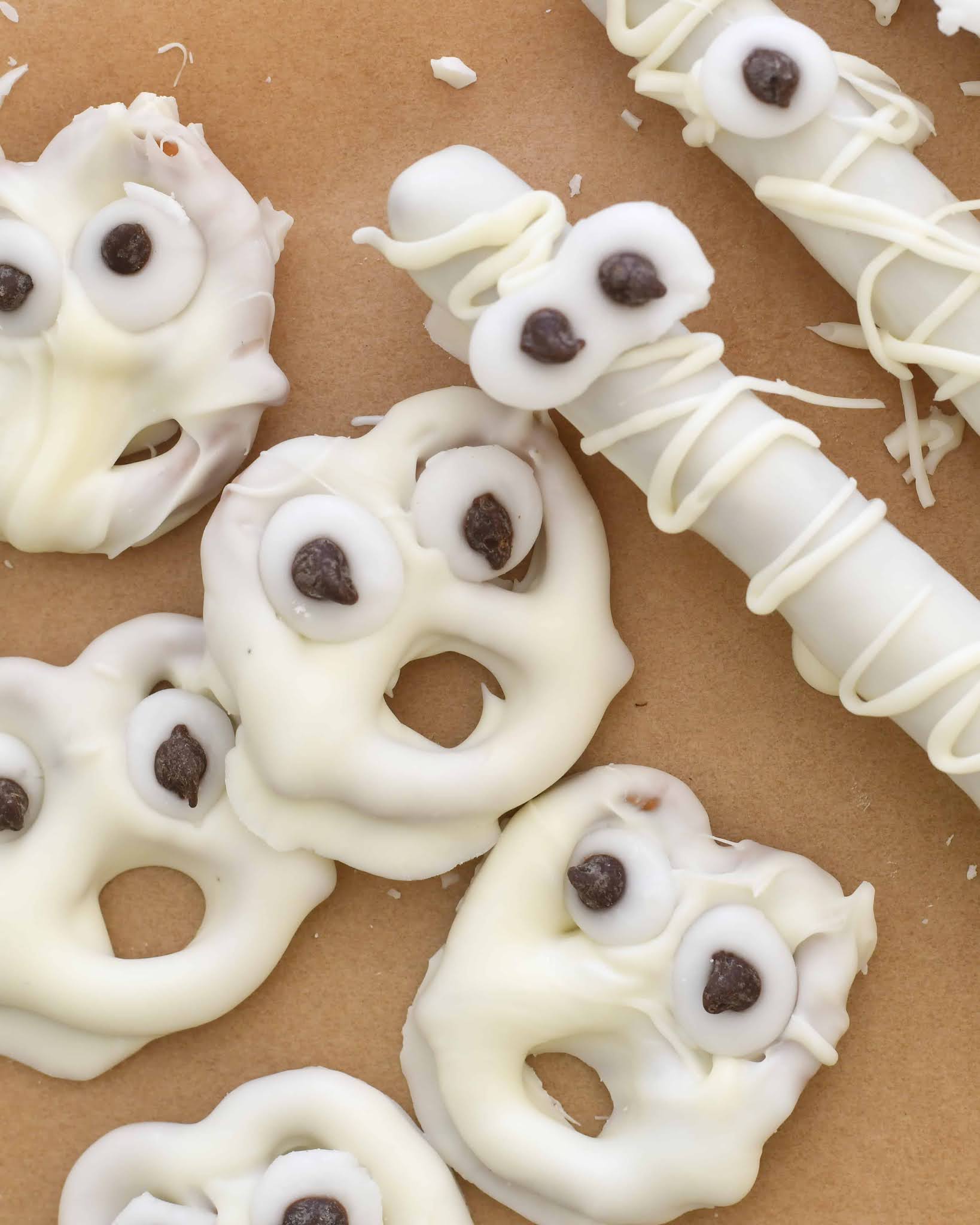 White chocolate-covered pretzels decorated to look like ghost faces with chocolate chip eyes and mouths, and a pretzel rod drizzled with white chocolate to resemble a mummy, all on brown parchment paper.