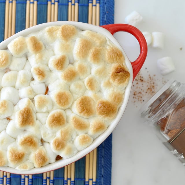 A red oval baking dish filled with toasted marshmallows sits on a blue placemat. An open jar of cinnamon and a few marshmallows are scattered nearby on a white surface.