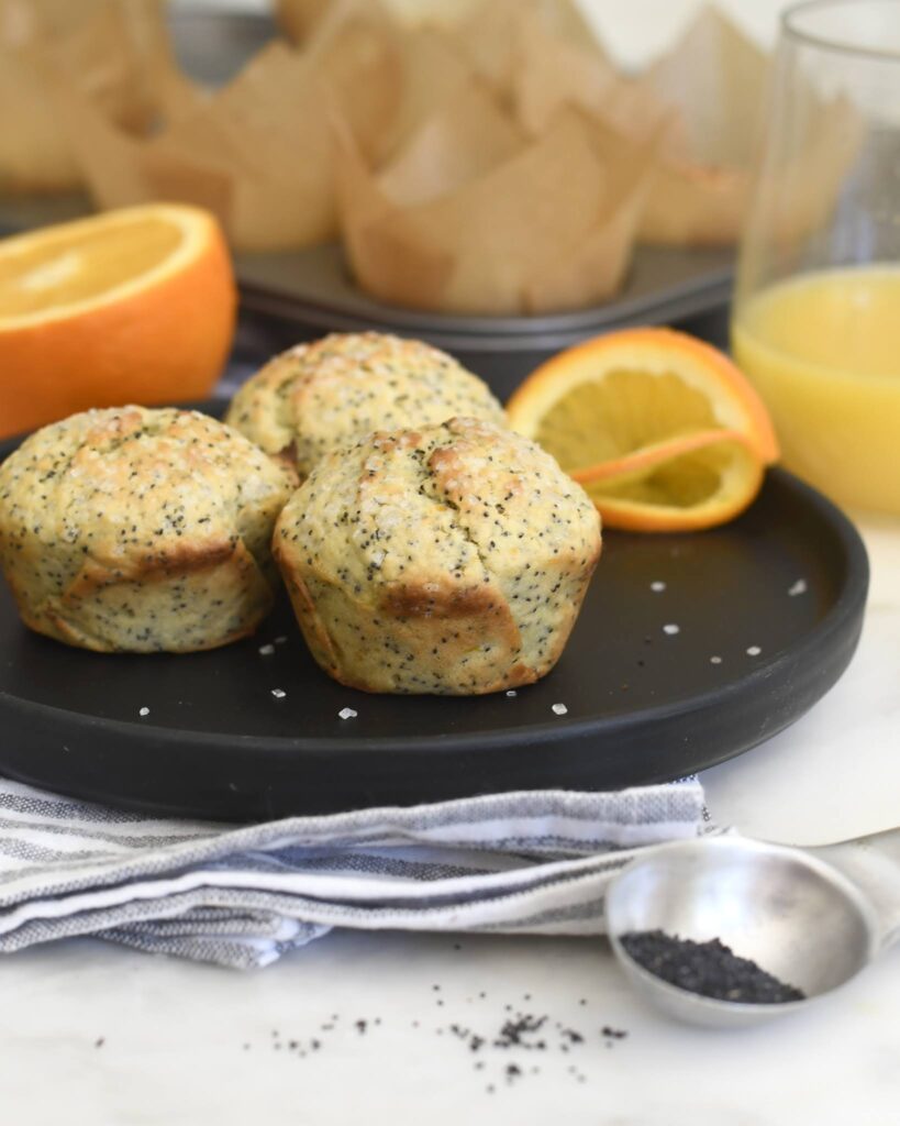 Three poppy seed muffins sit on a black plate with orange slices and a glass of orange juice. An orange, baking tray, and a measuring spoon with poppy seeds are in the background.