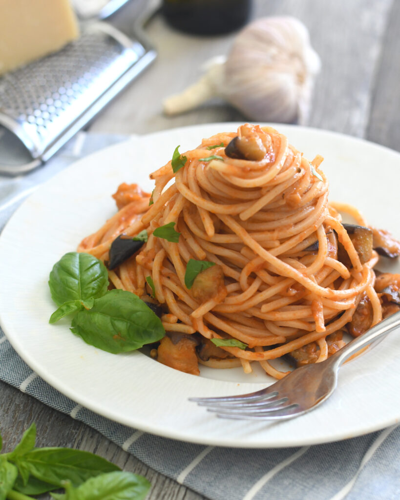 A neatly twirled mound of spaghetti with tomato sauce, small eggplant pieces, and fresh basil leaves is served on a white plate with a fork. Garlic, a cheese grater, and basil are in the background.