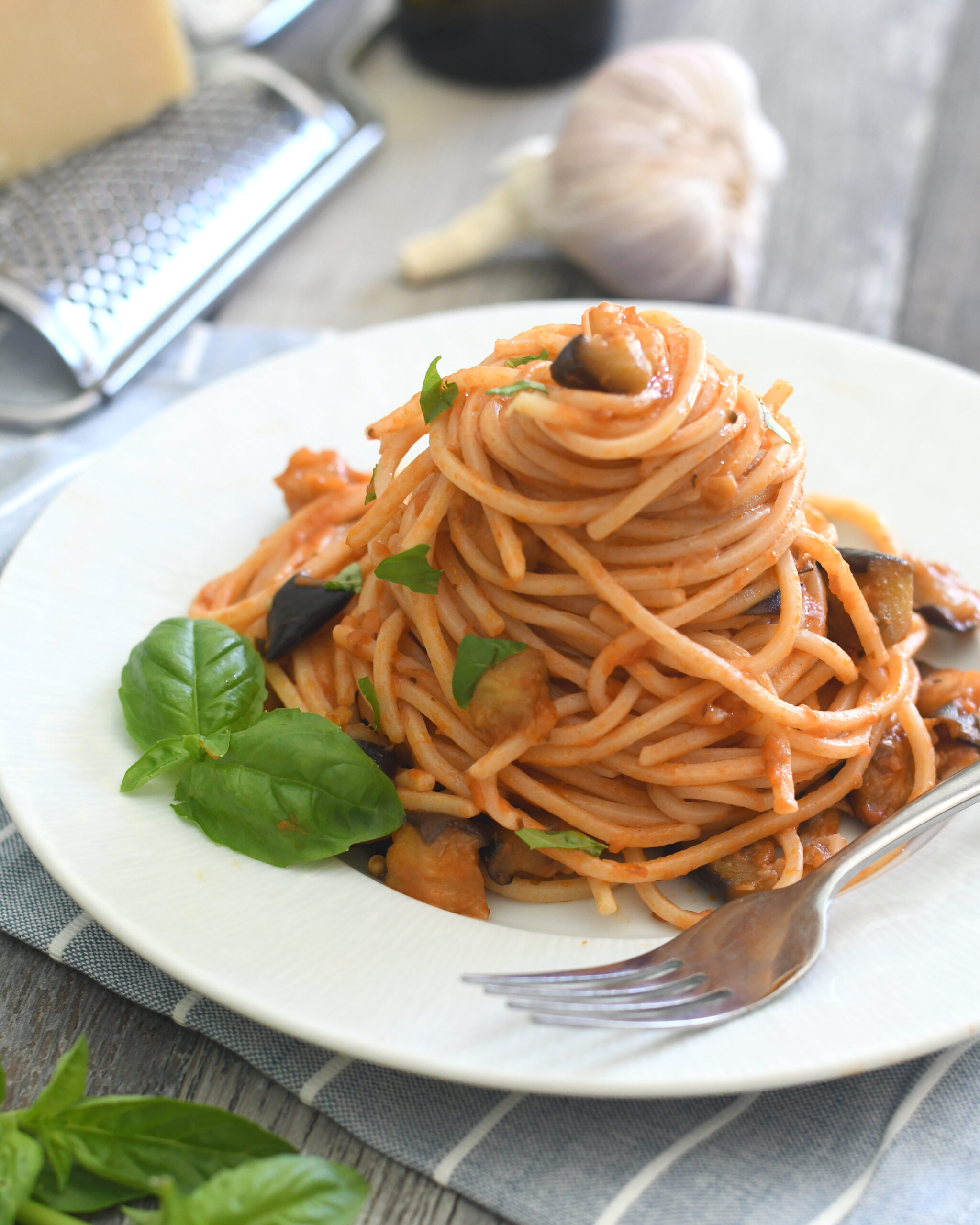 A neatly twirled mound of spaghetti with tomato sauce, small eggplant pieces, and fresh basil leaves is served on a white plate with a fork. Garlic, a cheese grater, and basil are in the background.