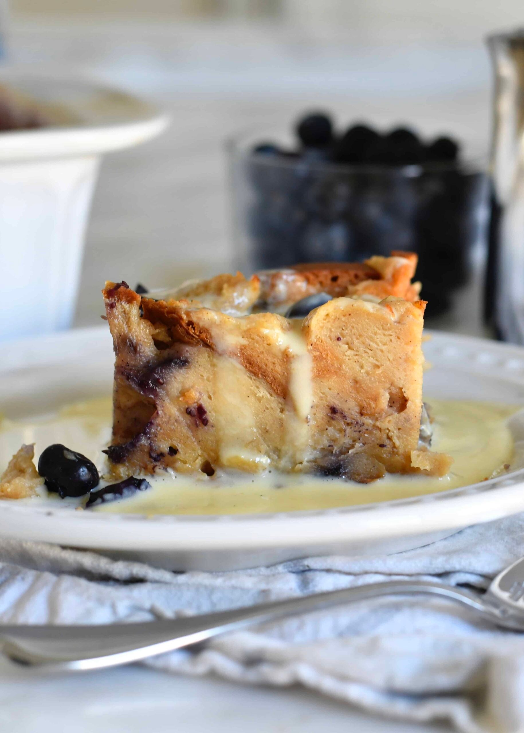A slice of bread pudding with blueberries sits on a white plate, topped with creamy vanilla sauce. Fresh blueberries and a jug are visible in the blurred background.