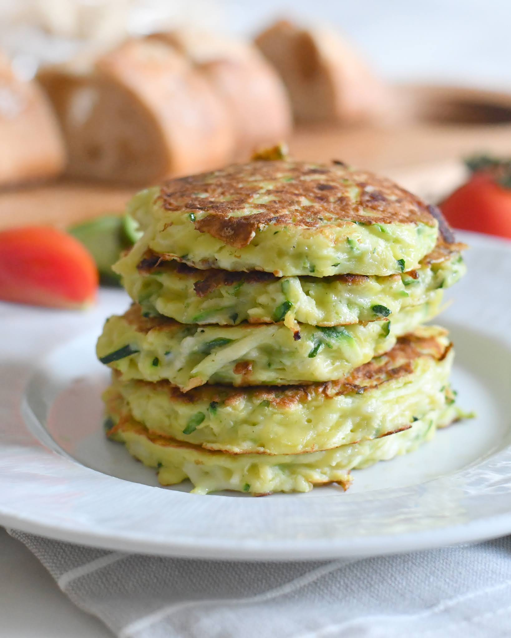 A stack of golden-brown zucchini fritters sits on a white plate, with fresh bread and tomatoes blurred in the background. The fritters are crisp on the outside and filled with visible green zucchini shreds.