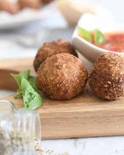 Three golden-brown arancini balls on a wooden board with fresh basil leaves, a jar of dried herbs, and a small white bowl of tomato dipping sauce in the background.