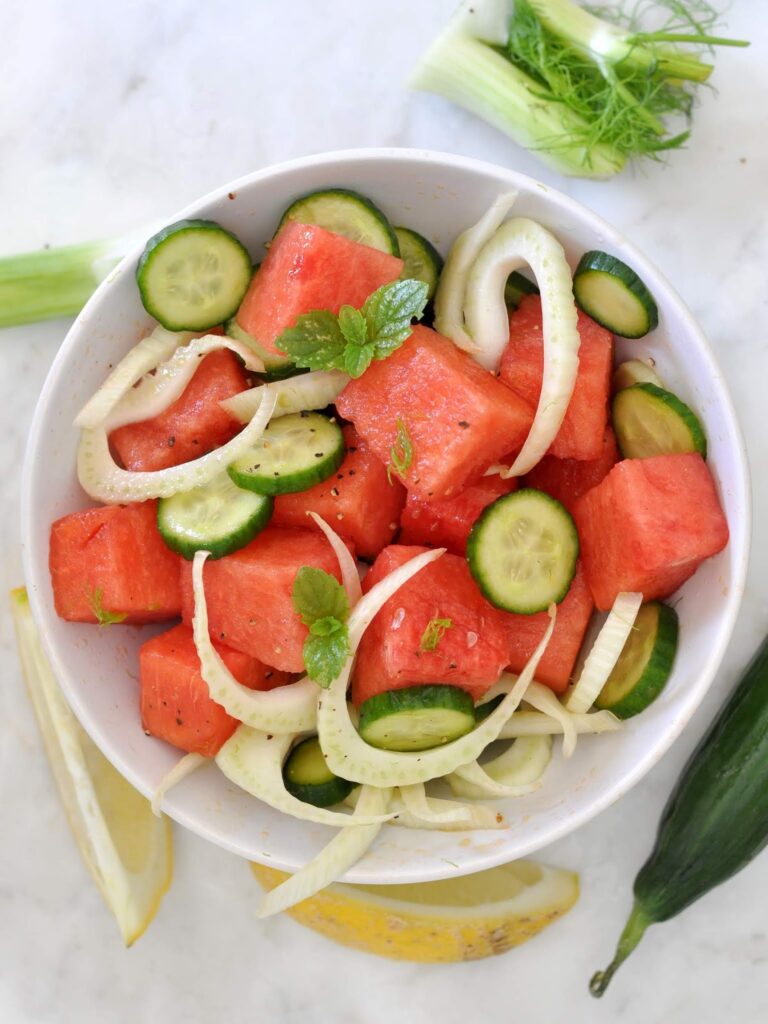 A bowl of salad containing watermelon cubes, cucumber slices, fennel, and mint leaves, garnished with black pepper. Cucumber, fennel, and lemon wedges are placed around the bowl on a white surface.
