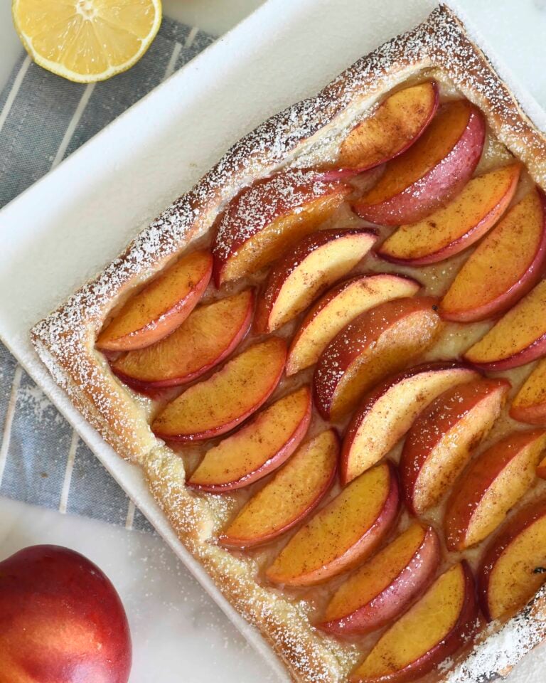 A rectangular peach tart with golden puff pastry, topped with sliced peaches and a dusting of powdered sugar sits on a white plate. A halved lemon and a whole peach are nearby on a striped cloth.