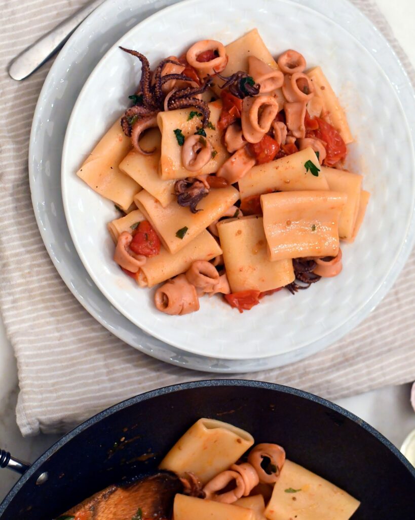 A white plate filled with pasta, tomato pieces, and calamari rings, garnished with chopped herbs, sits on a striped cloth next to a pan containing more of the same dish.