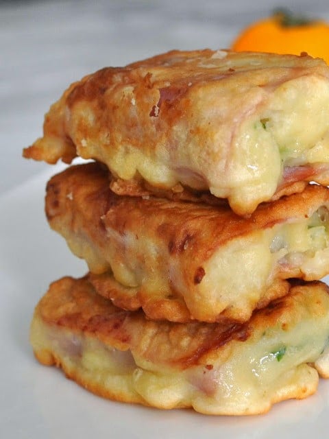 Three golden-brown, crispy fried sandwiches stacked on a plate, with melted cheese and bits of green and ham visible inside the batter. A blurred yellow fruit is in the background.