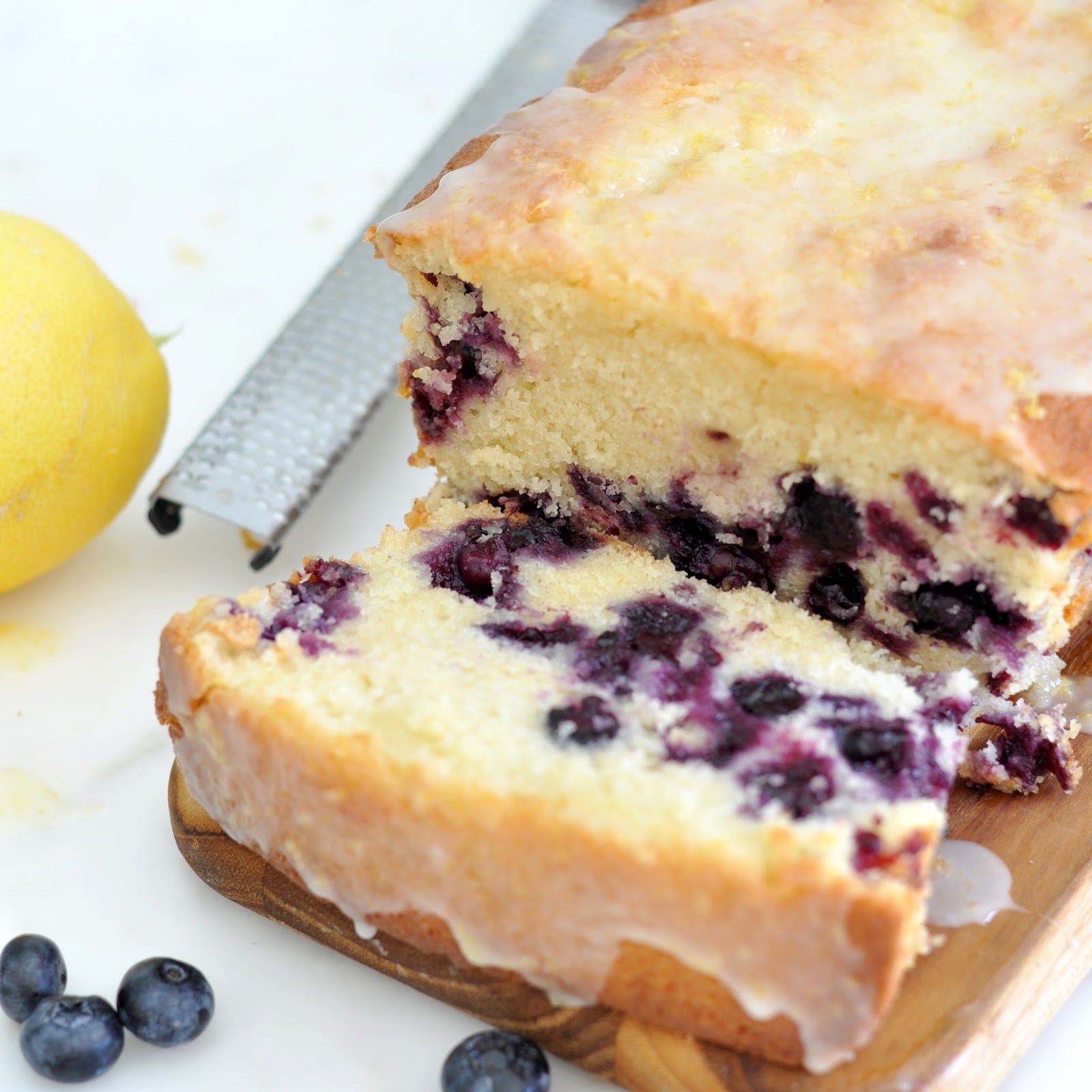 A sliced loaf of blueberry lemon bread with glaze, resting on a wooden board. Fresh blueberries and a lemon are beside the bread on a white surface, with a zester in the background.