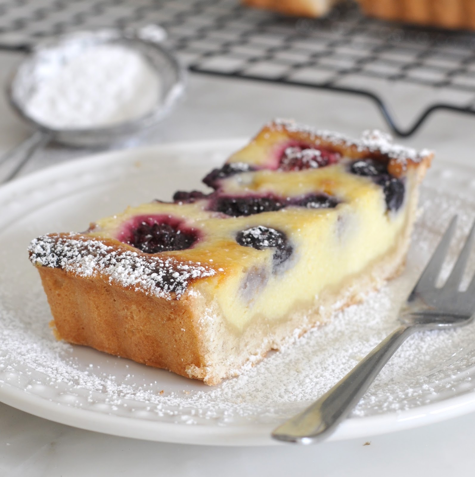 A slice of berry custard tart topped with powdered sugar sits on a white plate with a fork. More powdered sugar is sprinkled on the plate, and a sifter and cooling rack are in the blurred background.