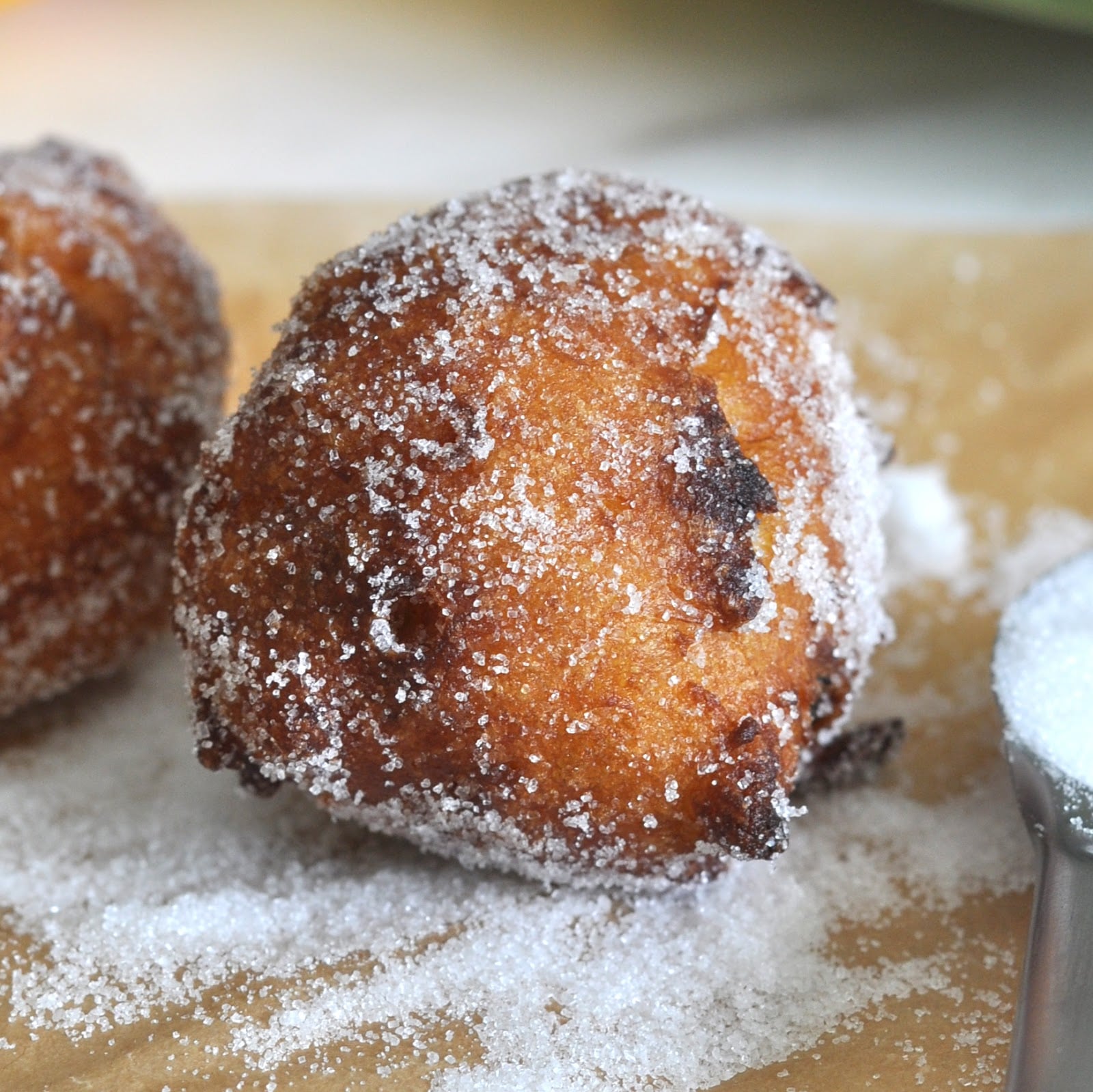 A close-up of a golden brown, round donut coated generously with granulated sugar, resting on a piece of parchment paper with more sugar scattered around.