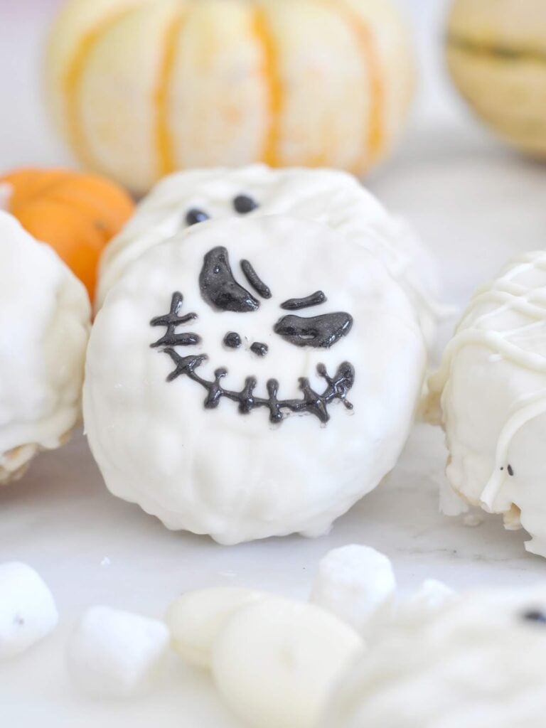 A white chocolate-covered treat decorated with a black Jack Skellington face sits on a white surface, with small marshmallows and white pumpkins in the background.