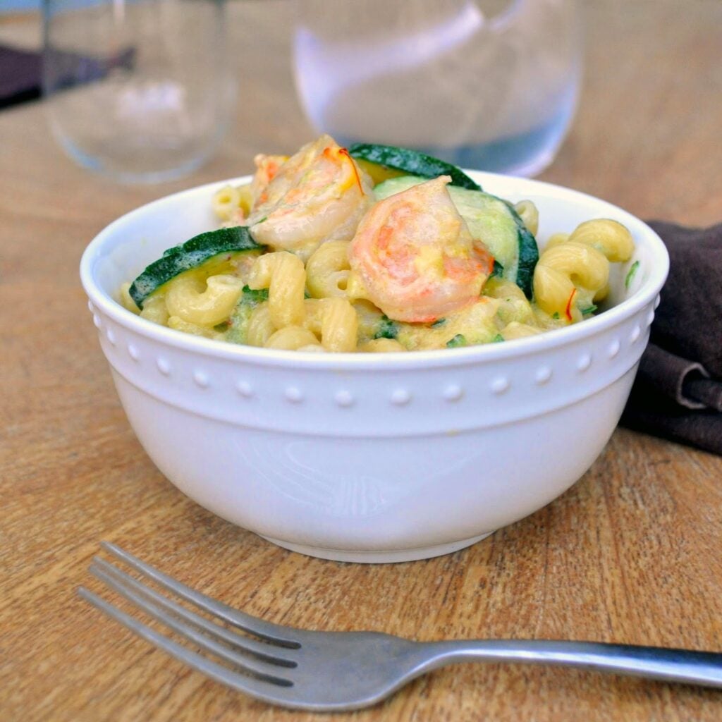 A white bowl filled with pasta, shrimp, and zucchini slices sits on a wooden table next to a fork and a brown napkin. A glass and a water pitcher are blurred in the background.