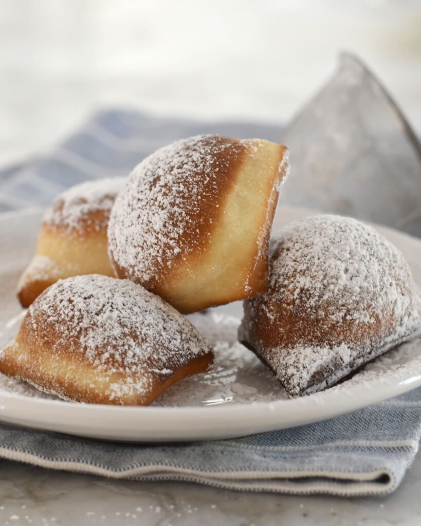 Three golden brown beignets dusted with powdered sugar sit on a white plate with a blue-striped napkin underneath. A metal sifter is blurred in the background.