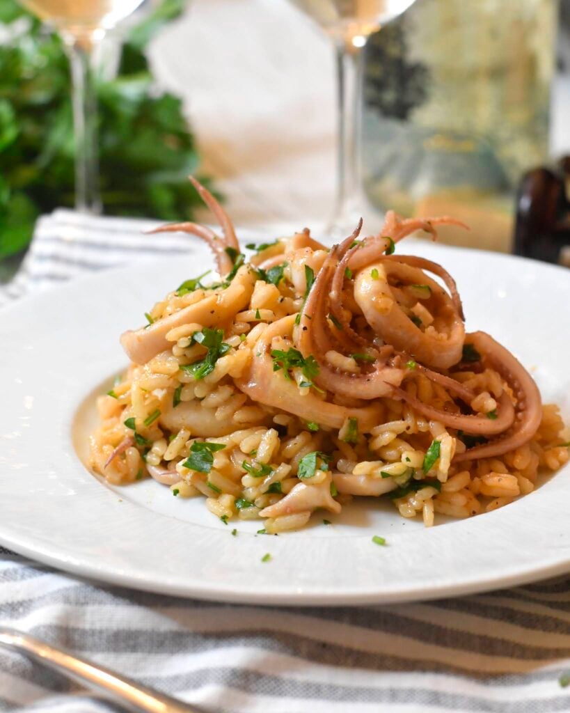 A plate of risotto topped with pieces of cooked squid and garnished with chopped parsley, served on a white plate with wine glasses and a wine bottle in the background.