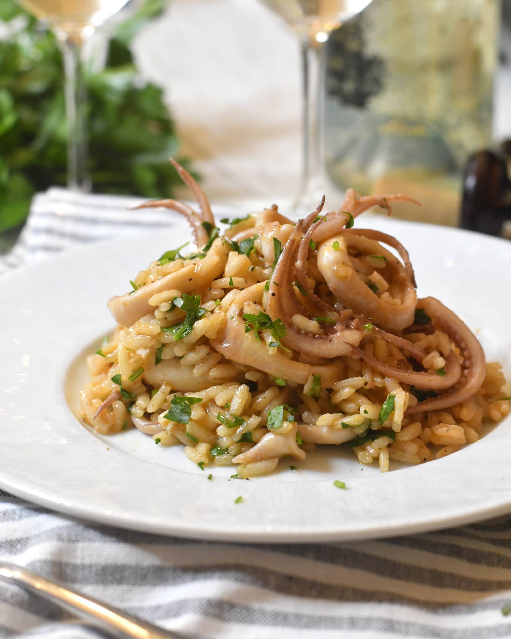 A white plate topped with risotto and pieces of squid, garnished with chopped parsley. The dish sits on a striped cloth, with wine glasses and a wine bottle blurred in the background.