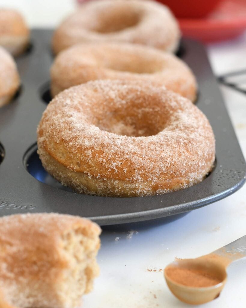 A close-up of cinnamon sugar donuts in a baking tray, with one donut in the foreground showing a bite taken out. A measuring spoon with cinnamon sugar sits nearby on a white surface.