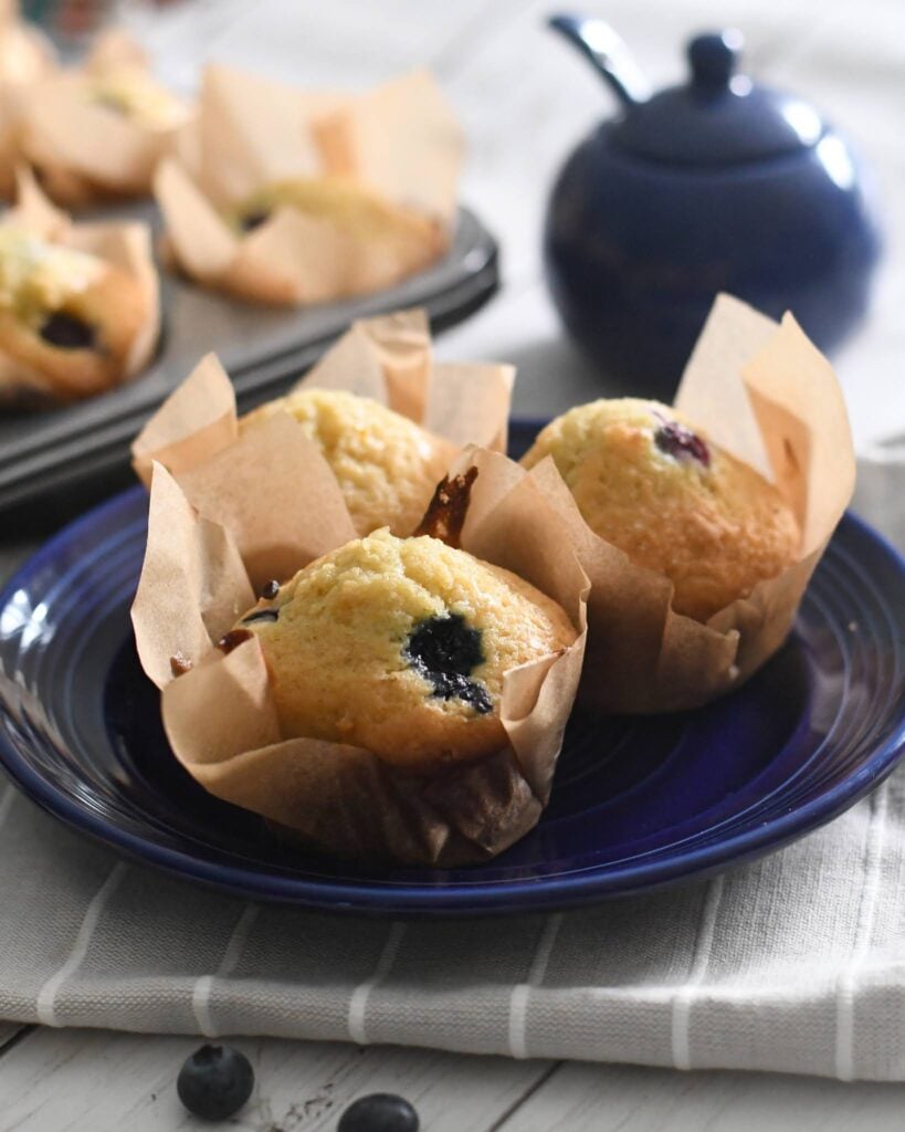 Three blueberry muffins in brown parchment paper on a dark blue plate, with a blue teapot and more muffins in a tray in the background. The scene is set on a striped cloth on a light wooden table.