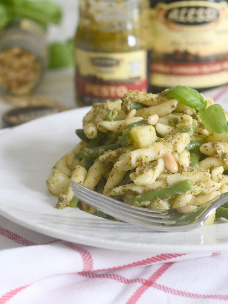 A plate of pasta with pesto sauce, green beans, zucchini, and pine nuts is garnished with fresh basil. A fork rests on the plate. Jars of pesto and another condiment are blurred in the background.