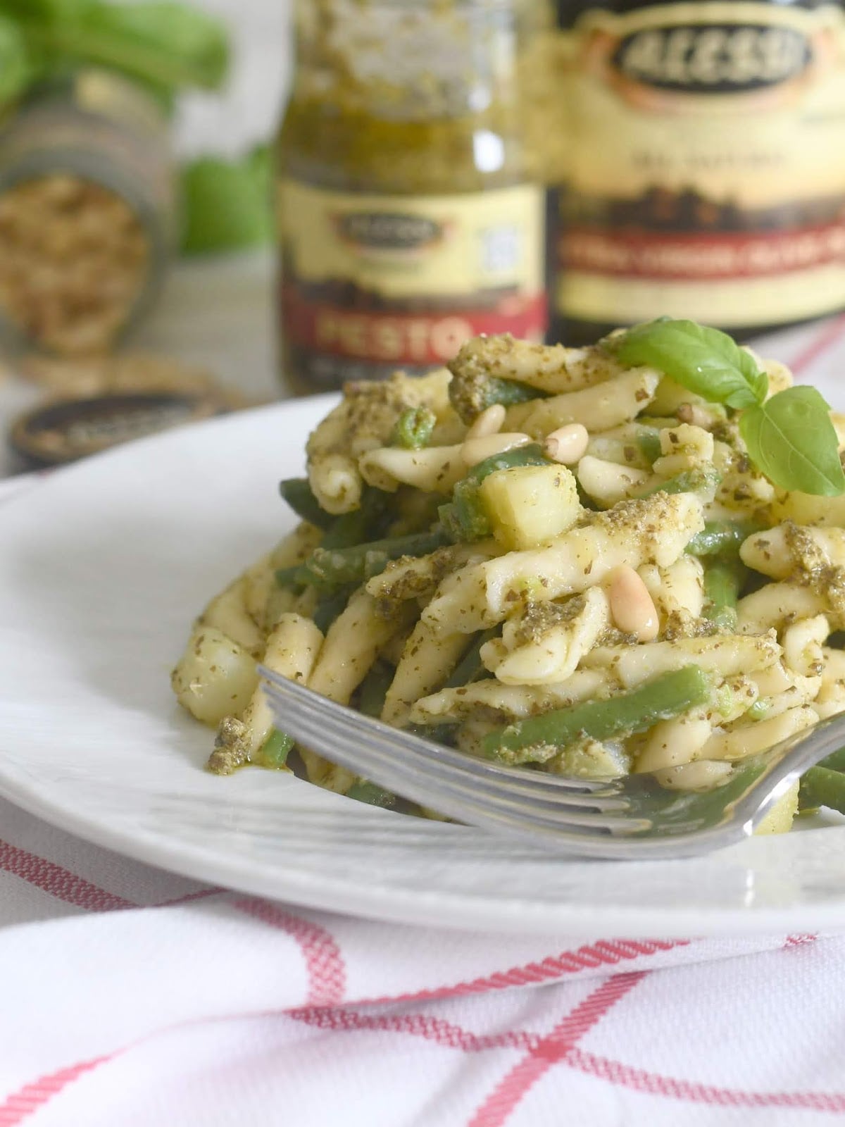 A plate of pasta with pesto sauce, green beans, zucchini, and pine nuts is garnished with fresh basil. A fork rests on the plate. Jars of pesto and another condiment are blurred in the background.
