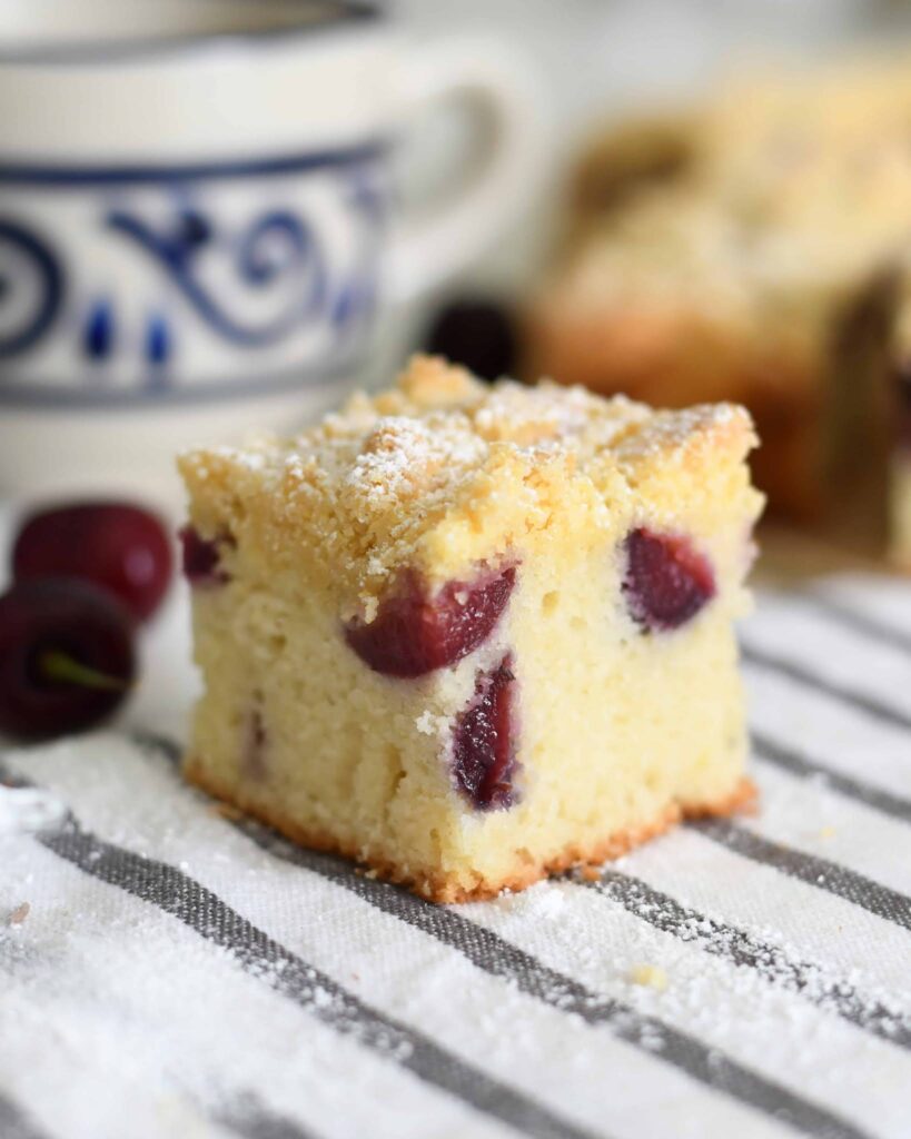 A square slice of cherry cake with a light dusting of powdered sugar sits on a striped cloth, with fresh cherries and a patterned mug in the softly blurred background.