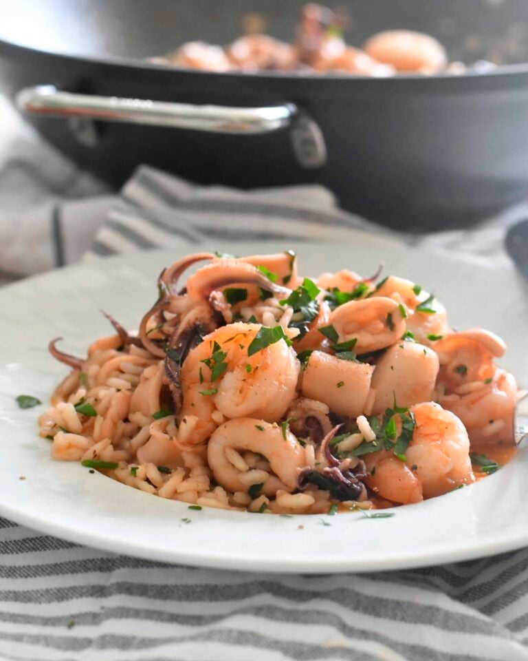 A plate of seafood risotto with shrimp, squid, and fresh parsley sits on a striped napkin. A black skillet with more seafood is visible in the blurred background.