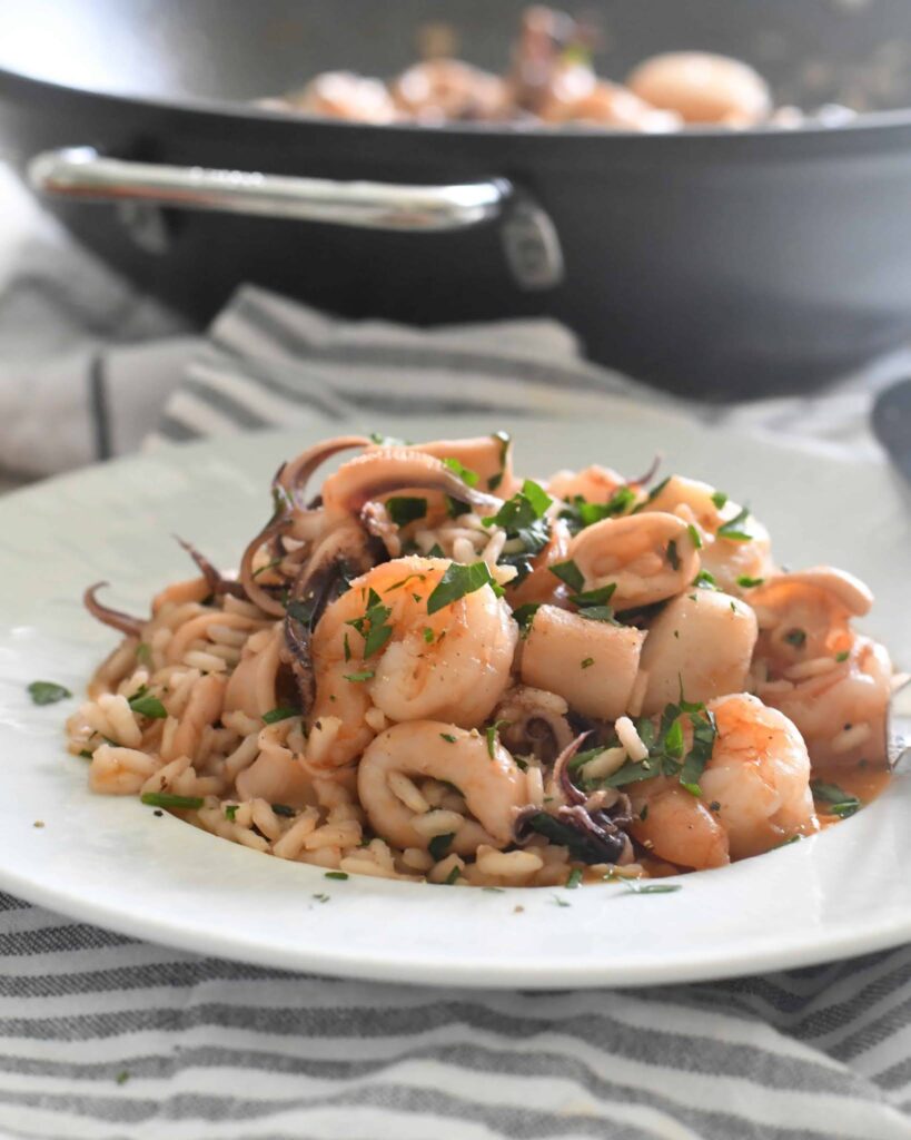 A plate of seafood risotto with shrimp, squid, and fresh parsley sits on a striped napkin. A black skillet with more seafood is visible in the blurred background.