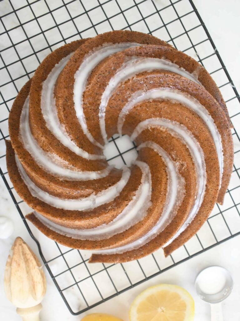 A spiral-patterned bundt cake drizzled with white glaze sits on a black cooling rack, surrounded by a wooden citrus reamer, a metal measuring spoon, and a sliced lemon on a white surface.