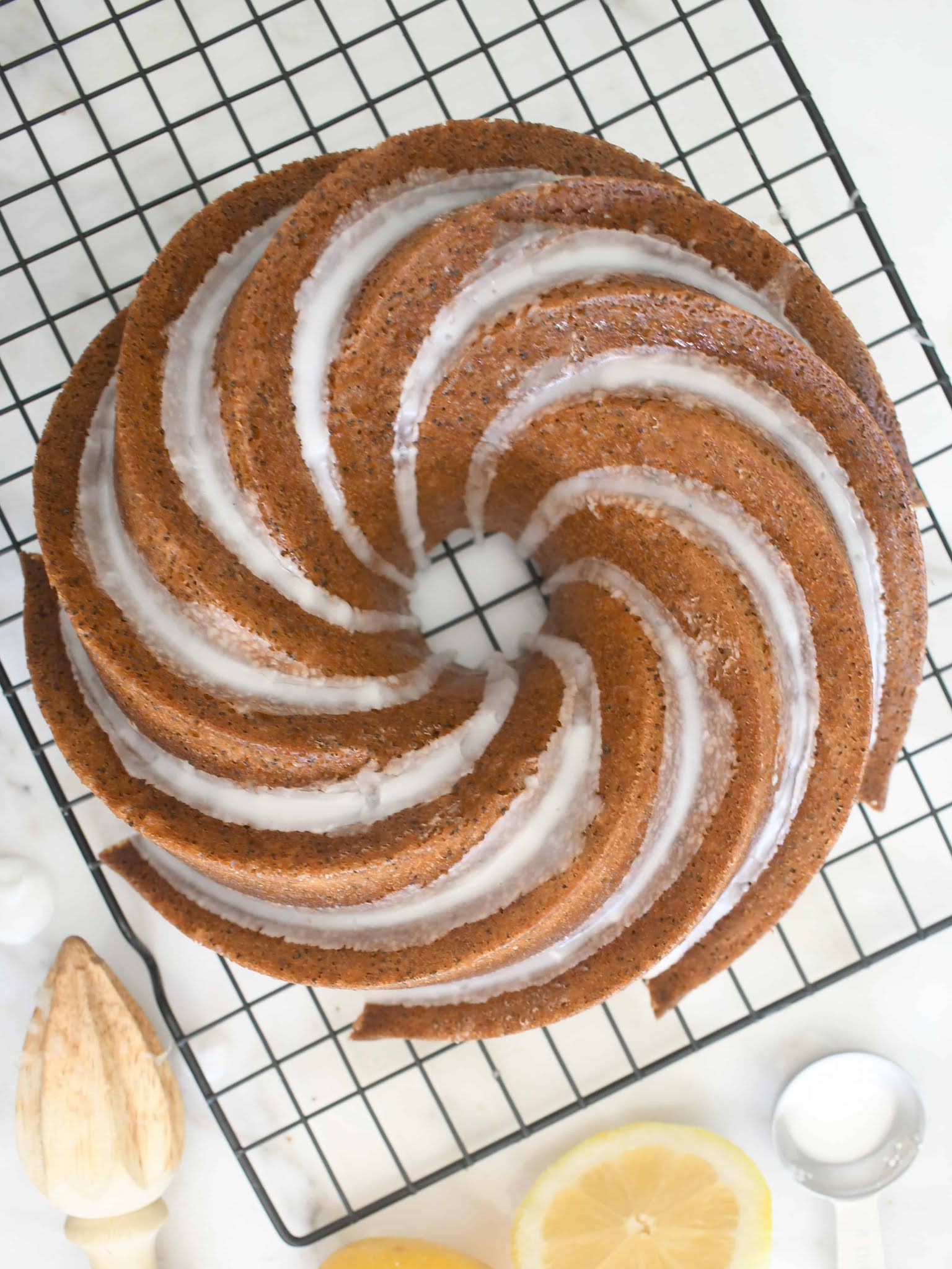 A spiral-patterned bundt cake drizzled with white glaze sits on a black cooling rack, surrounded by a wooden citrus reamer, a metal measuring spoon, and a sliced lemon on a white surface.