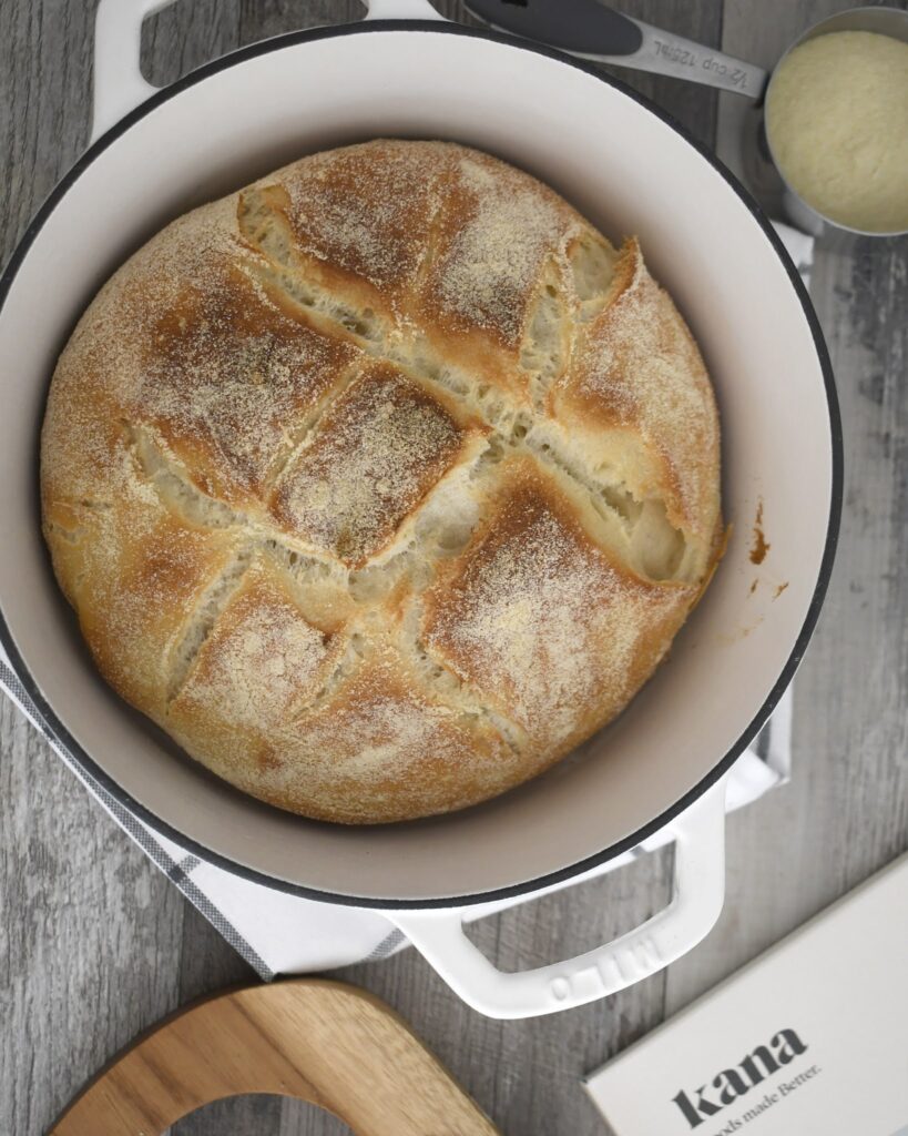 A round loaf of rustic bread with a golden crust sits in a white Dutch oven on a wooden surface, surrounded by a wooden board, a book titled kana, and some kitchen utensils.