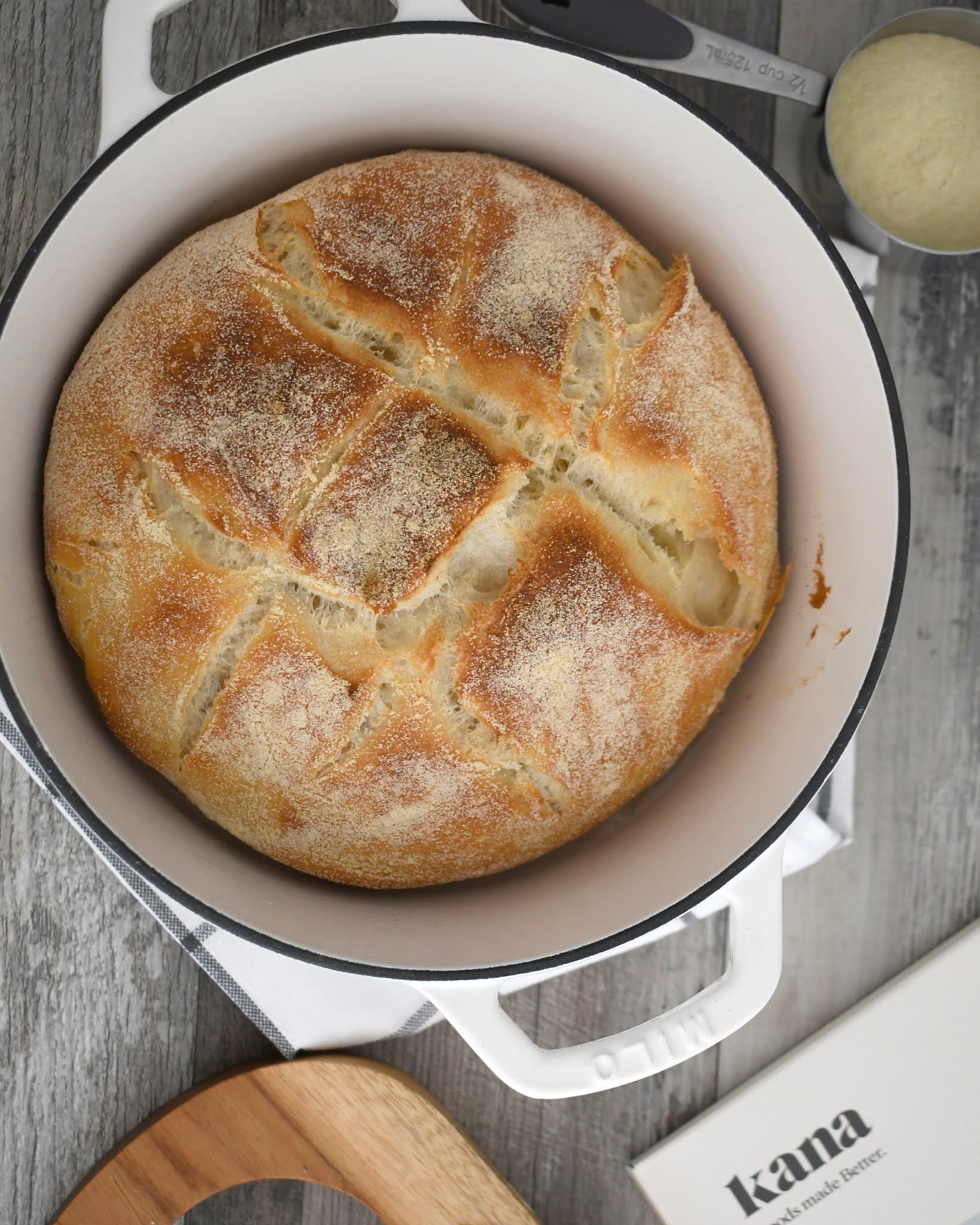 A round loaf of rustic bread with a golden crust sits in a white Dutch oven on a wooden surface, surrounded by a wooden board, a book titled kana, and some kitchen utensils.
