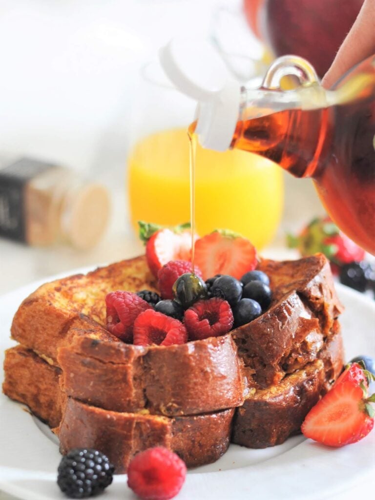 Thick slices of French toast topped with fresh strawberries, raspberries, blueberries, and blackberries, with syrup being poured over them. A glass of orange juice is visible in the background.