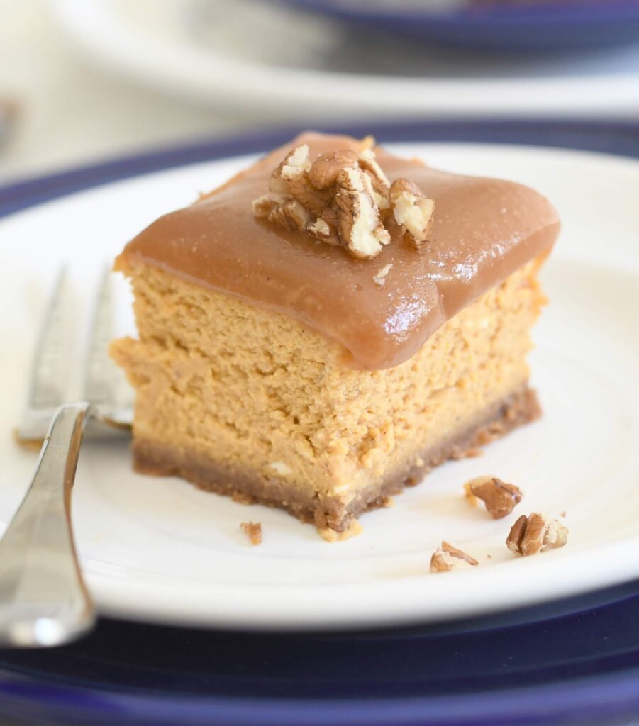 A square slice of cheesecake with a crumb crust, topped with a thick layer of caramel and chopped pecans, sits on a white plate with a fork beside it.