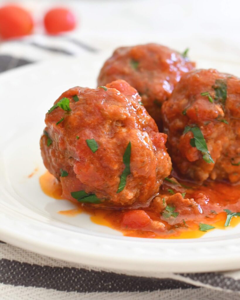 Three meatballs topped with tomato sauce and garnished with chopped parsley, served on a white plate. Some sauce and herbs are visible on the plate. The background is softly blurred.