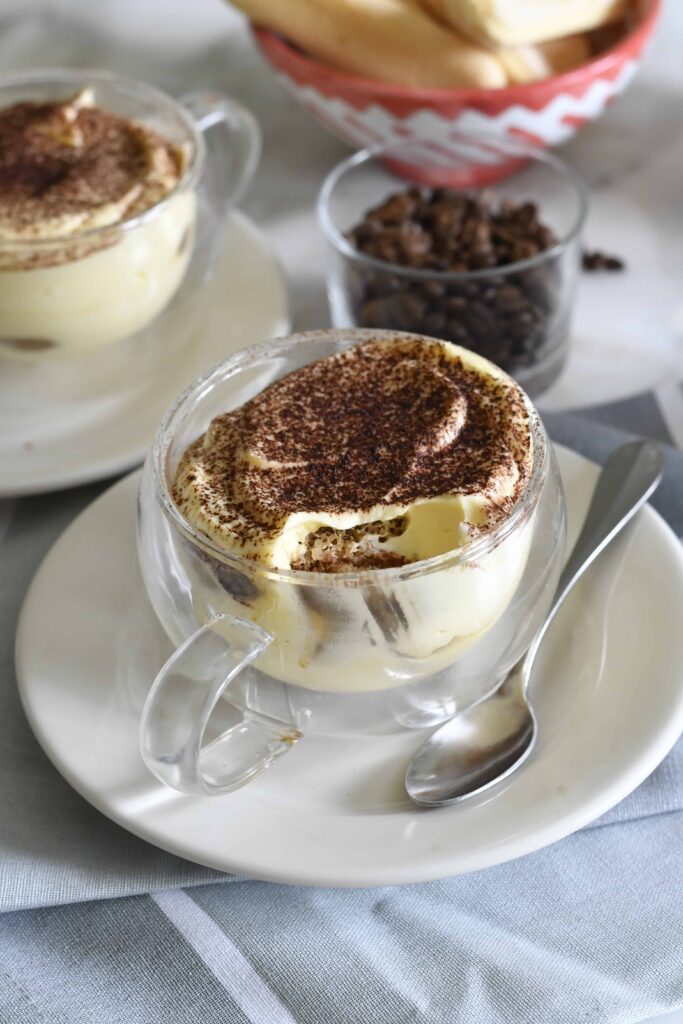 A glass cup filled with tiramisu, topped with cocoa powder, sits on a white plate with a spoon beside it. In the background, another cup of tiramisu, coffee beans, and ladyfingers are visible.