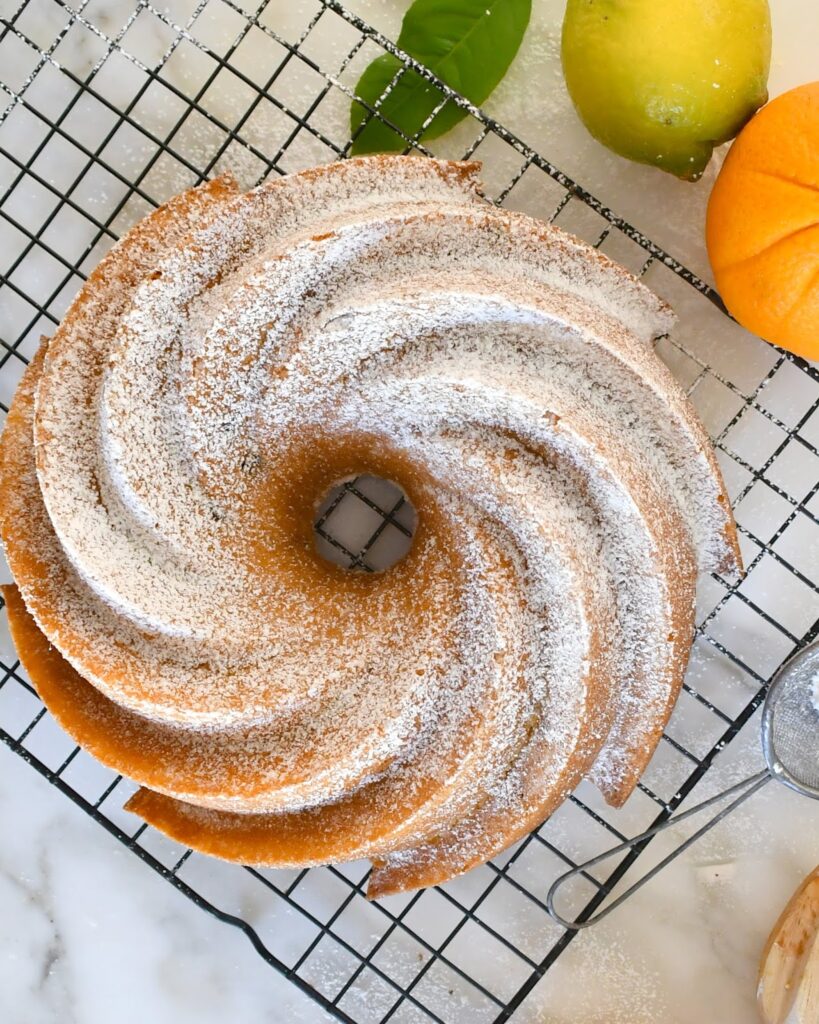 A golden bundt cake dusted with powdered sugar sits on a wire cooling rack. Fresh lemons and leaves are nearby, and a sifter rests on the marble countertop.
