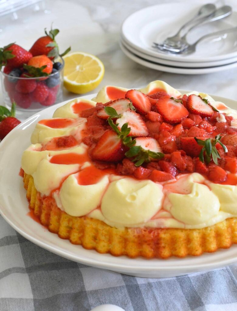 A sponge cake topped with creamy custard and fresh strawberries, drizzled with strawberry sauce, sits on a white plate beside a container of strawberries, a lemon, and stacked white plates with forks.