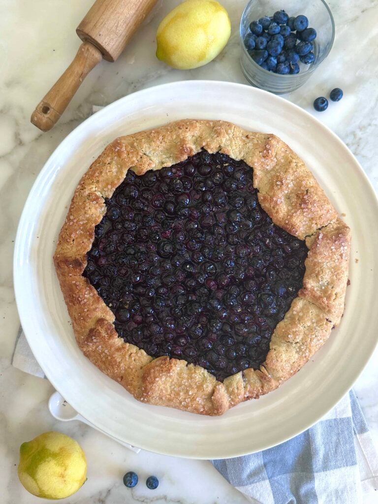 A rustic blueberry galette with a golden, sugar-dusted crust sits on a white plate, surrounded by lemons, a bowl of fresh blueberries, and a wooden rolling pin on a marble surface.