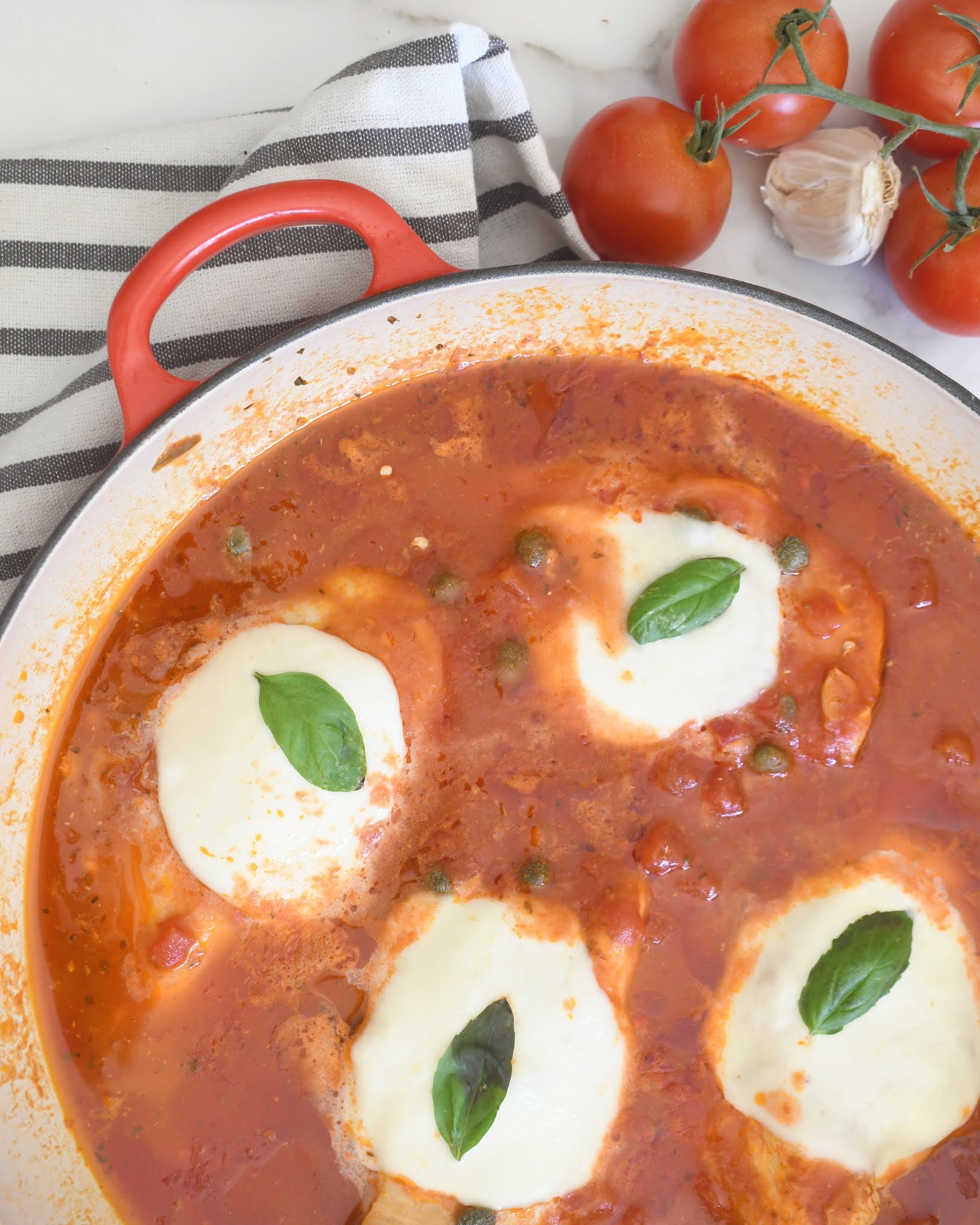 A pan of tomato sauce with four cheese-topped baked eggs, each garnished with a basil leaf. A striped towel, fresh tomatoes on the vine, and garlic are beside the pan.
