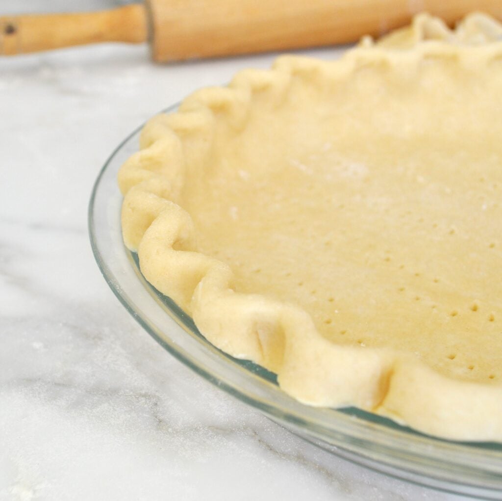 A glass pie dish filled with unbaked pie crust, edges crimped, and small fork holes in the base. A wooden rolling pin is visible in the background on a marble surface.